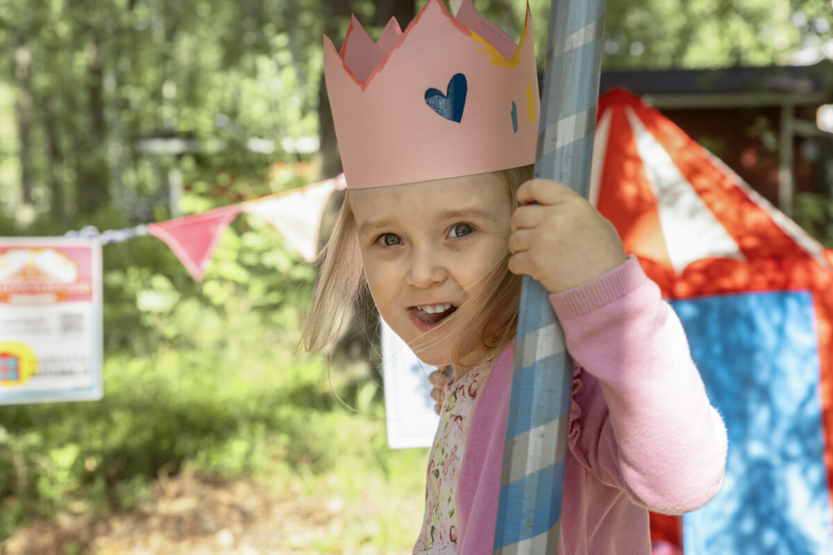 Iloinen lapsi vaaleanpunainen pahvikruunu päässään. Taustalla metsää ja värikkäitä viirejä.